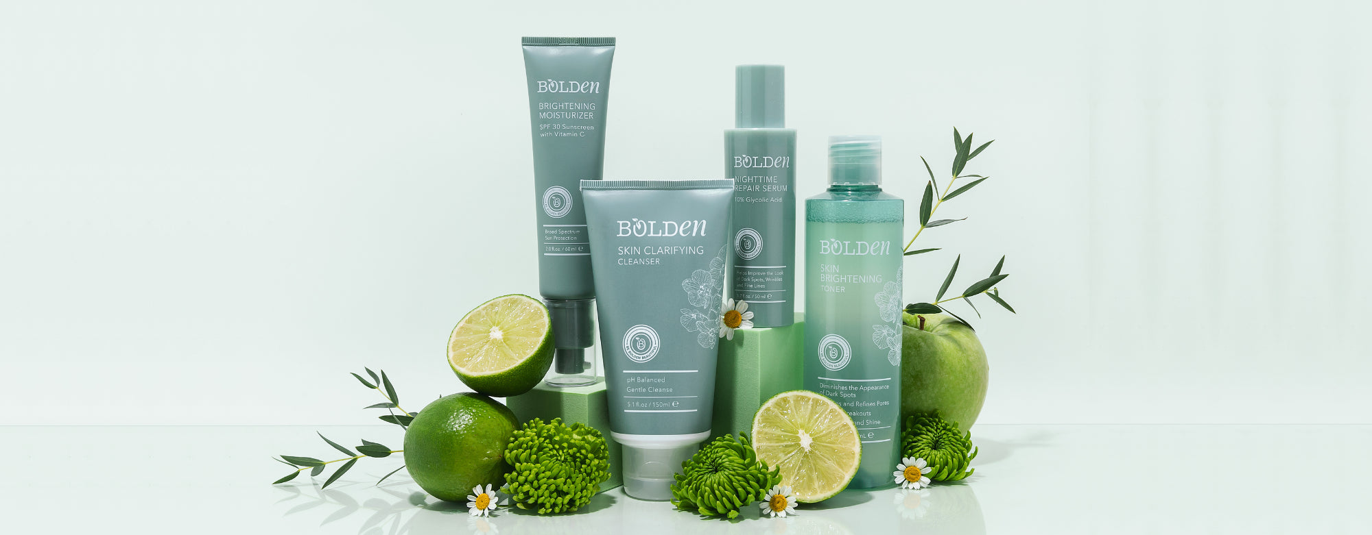  Four Bolden skincare products stand among an assortment of greenery, limes, and an apple. From left to right: Brightening Moisturizer, Skin Clarifying Cleanser, Nighttime Repair Serum, Skin Brightening Toner.