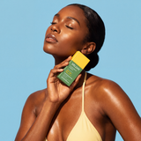 A woman in a pale yellow bikini top holding Bolden sunscreen serum bottle against a solid light blue background.