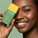 A smiling woman holds a Bolden Sunscreen Serum box next to her face against a soft green background. The box is green with a yellow top and white text, showing SPF 50+ PA++++ protection.
