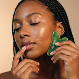 Woman using a green facial tool on her face against a beige background