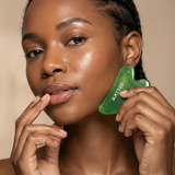Woman using a green jade gua sha on her face against a beige background