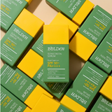 A top-down flat lay of Bolden sunscreen serum boxes, with one central green and yellow box standing upright while several matching boxes surround it on a light beige surface.